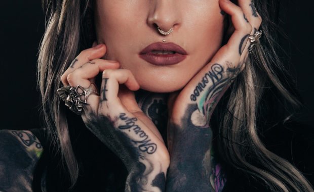 Woman with blonde hair & nose ring with tattoos hands & arms holding face - summer tattoo